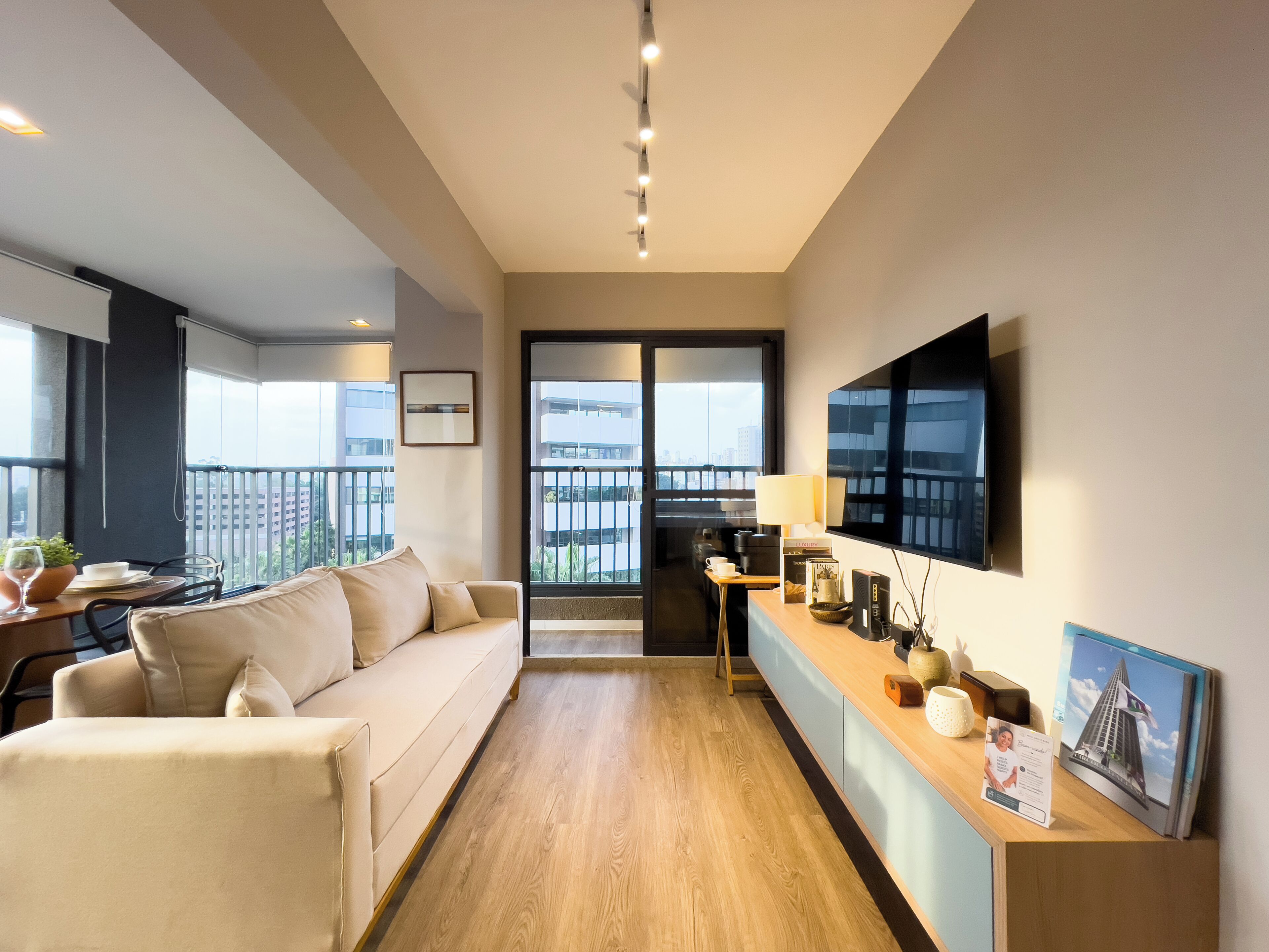A modern living room with a sofa, a coffee table, and a TV. There is a balcony with a view of buildings.
