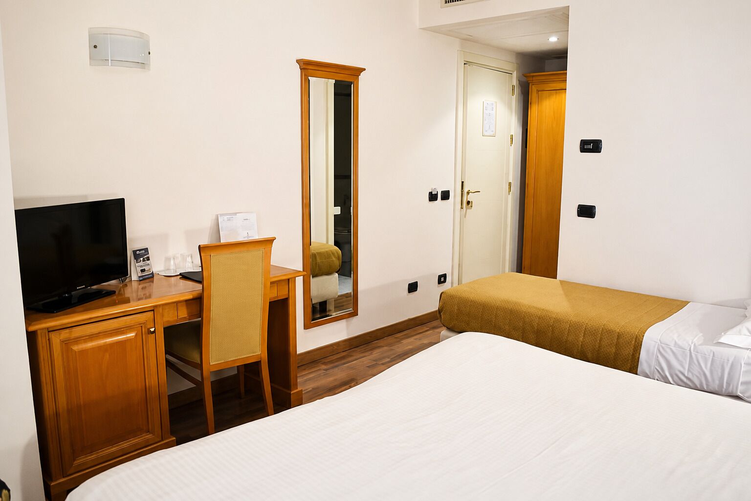 Ein Hotelzimmer mit einem Bett, einem Schreibtisch mit Fernseher, einem Sessel, einem Spiegel und einem Kleiderschrank.