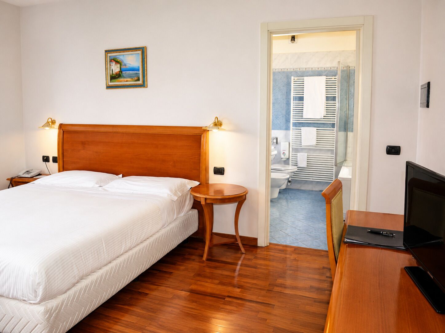 Una camera d'albergo con un letto di legno, lampade ai comodini, una scrivania con un computer e vista sul balcone.