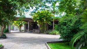 A paved pathway leading to a building with a covered entrance, surrounded by lush greenery and palm trees.