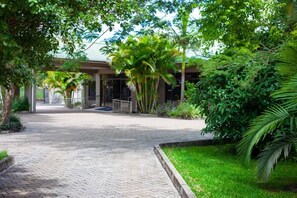 Un sendero pavimentado conduce a un edificio con acceso cubierto, rodeado de exuberante follaje y palmeras.