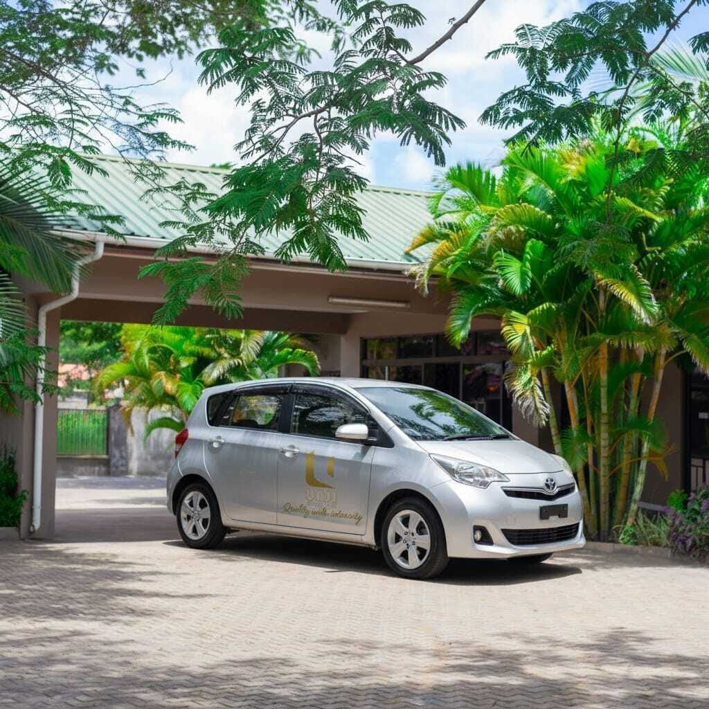 Um carro Toyota prata estacionado em frente a um edifício com entrada coberta e plantas tropicais.
