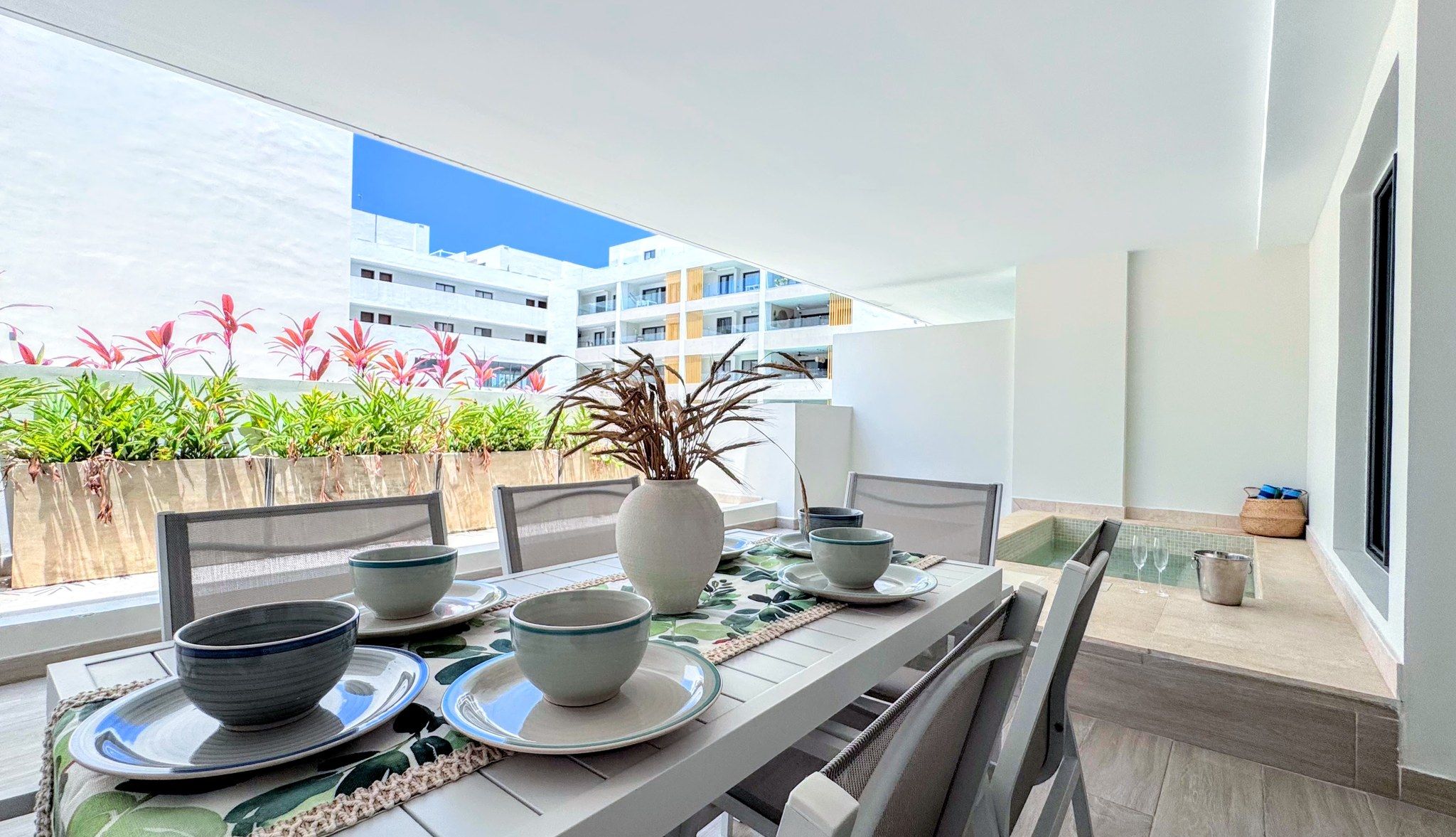 A dining area with a table set for four, chairs, a vase with dried plants, and a view of a building with balconies.