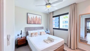 Ein Hotelzimmer mit einem Bett, einem Nachttisch, einem Deckenventilator, einem Fenster mit Vorhängen und Blick auf Gebäude.