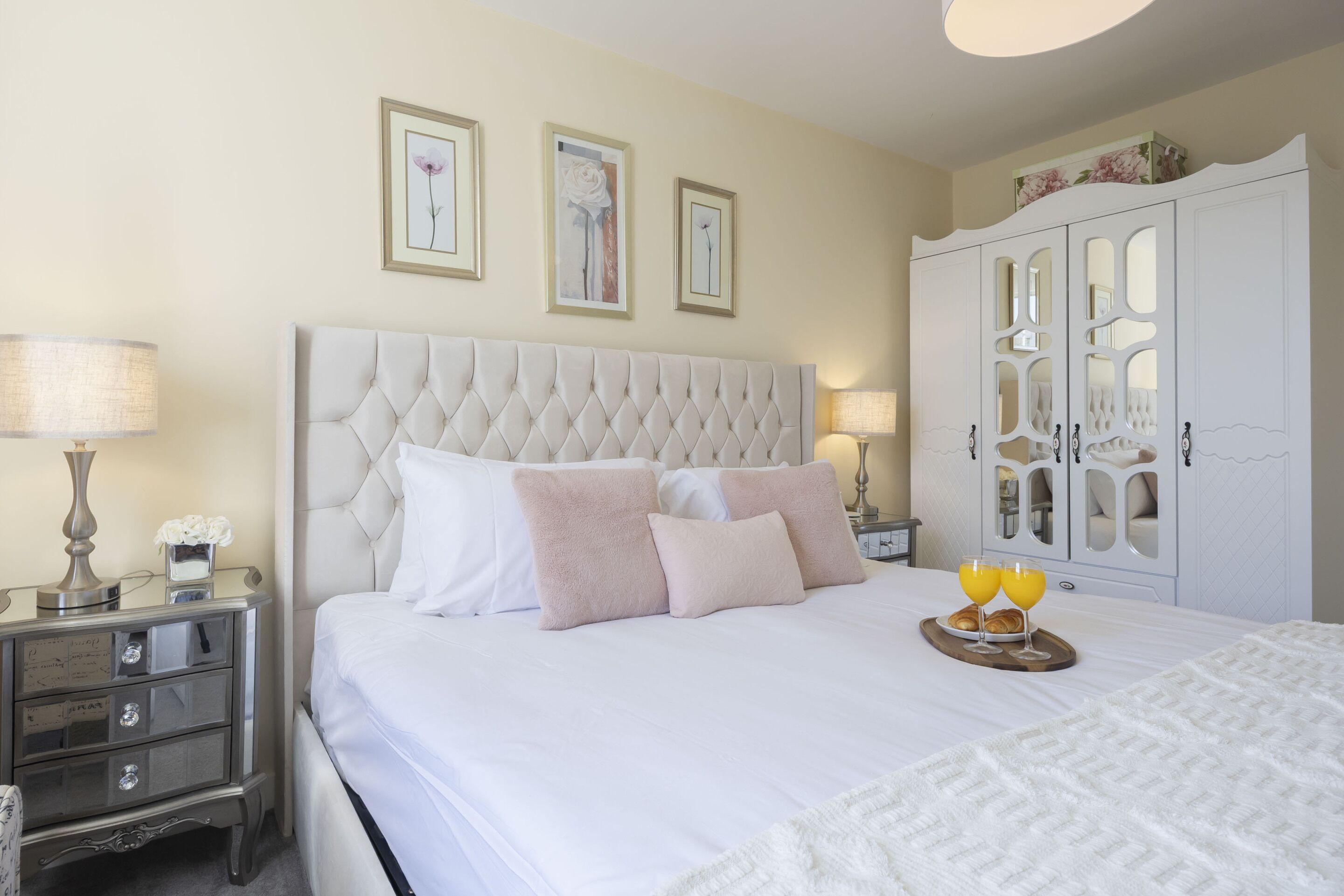 A neatly made bed with a tufted headboard, two bedside lamps, a nightstand with a mirror, and a cabinet with mirrored doors.