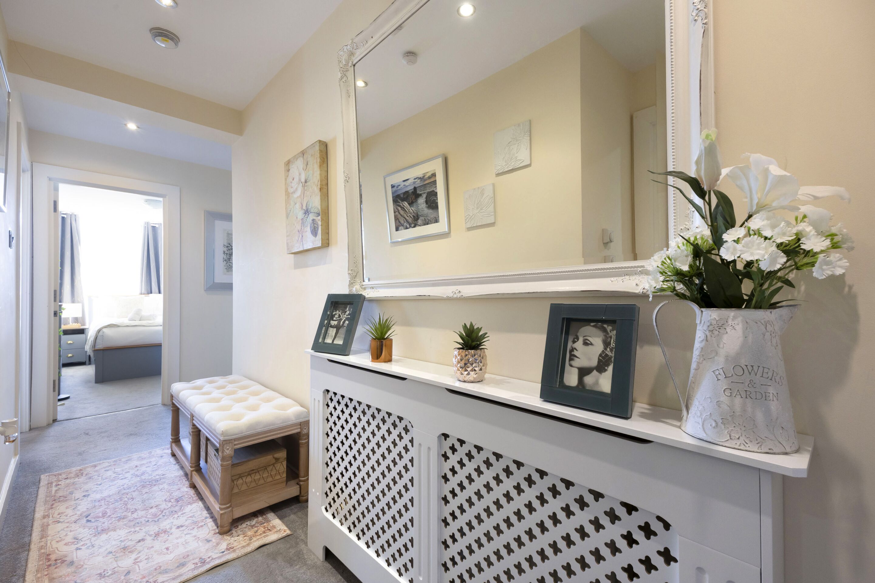 Un couloir avec une console blanche, un banc et un grand miroir. On y trouve des tableaux encadrés et un vase avec des fleurs posé sur la console.