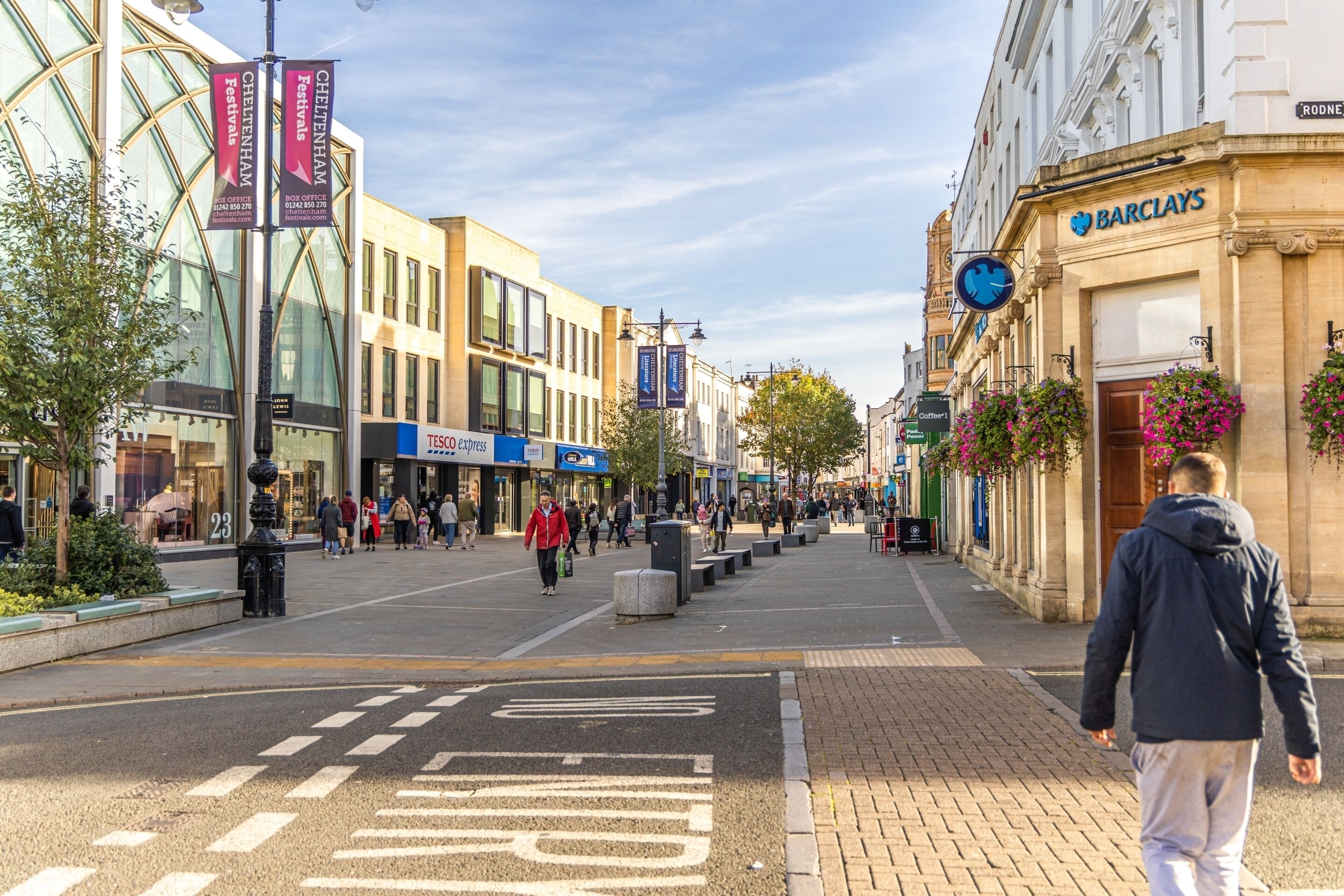 Uma rua destinada a pedestres, repleta de lojas e cafés, incluindo um banco Barclays e uma loja Tesco Express.