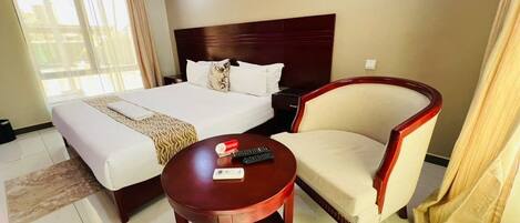 Quarto de hotel com cama, uma cadeira, uma mesa pequena e uma janela com cortinas.