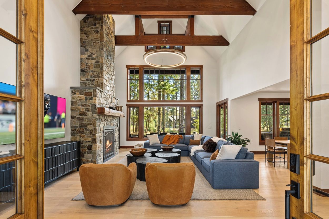 A spacious living room with a stone fireplace, large windows, and a high ceiling with exposed beams.