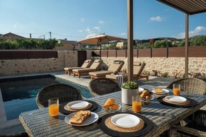 Un espace repas aménagé au bord de la piscine, avec des meubles en osier, une table dressée pour le petit-déjeuner et une vue sur le paysage environnant.
