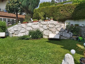 Een stenen muur met een bankje, een kleine tuin met gras en planten, en een gebouw op de achtergrond.