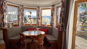 A cozy corner seating area with a round table, cushioned benches, and a view of a snowy landscape through large windows. - Apartment Sibylle by Alpine Host Helpers (Kirchberg in Tirol)