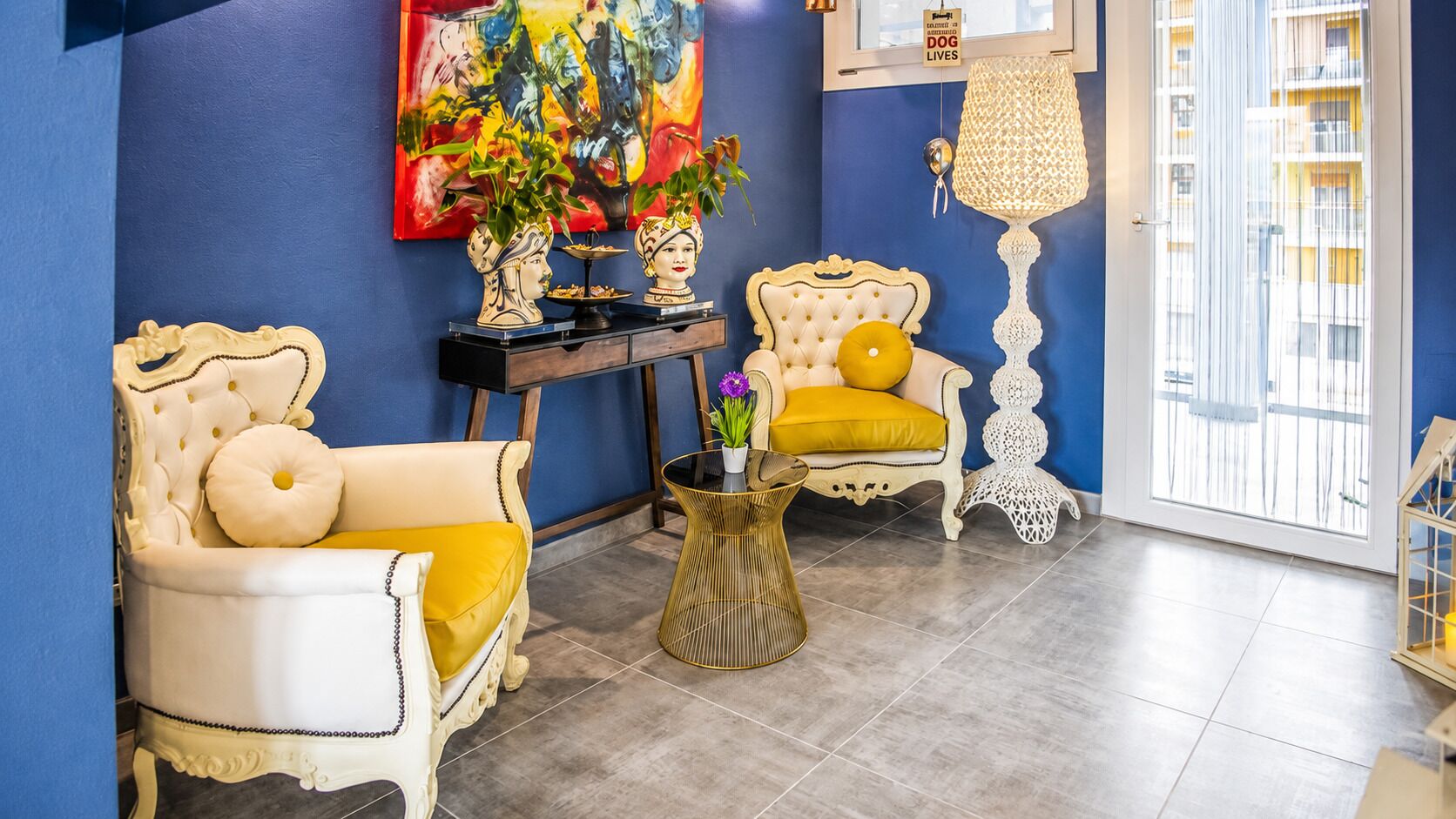 A room with two white armchairs with yellow cushions, a small round table with a vase, a painting on the wall, and a window with blinds.