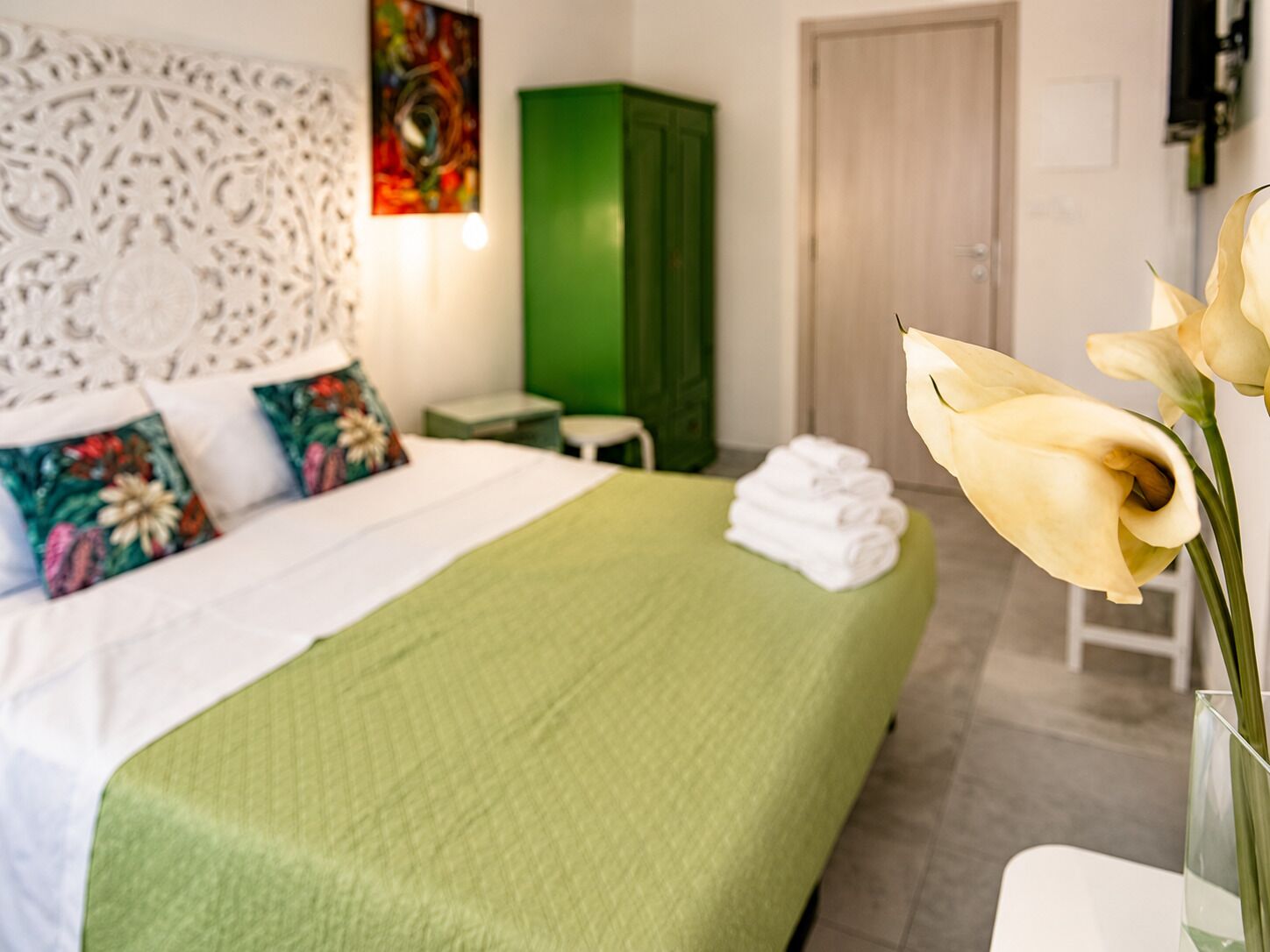 A hotel room with a bed, pillows, a green cabinet, a white door, and a vase with flowers.
