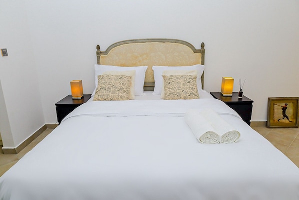 A neatly made bed with white linens, two pillows with floral patterns, and a rolled towel on the bed. There are two bedside tables with lamps and a framed picture on one.