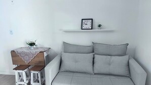 Ein modernes Wohnzimmer mit einem grauen Sofa, zwei weißen Sesseln, einem Holztisch mit Spitzenbettdecke, einer kleinen Topfpflanze und einer an der Wand befestigten Uhr.