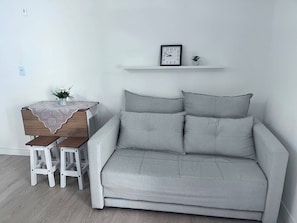 Ein modernes Wohnzimmer mit einem grauen Sofa, zwei weißen Sesseln, einem Holztisch mit Spitzenbettdecke, einer kleinen Topfpflanze und einer an der Wand befestigten Uhr.