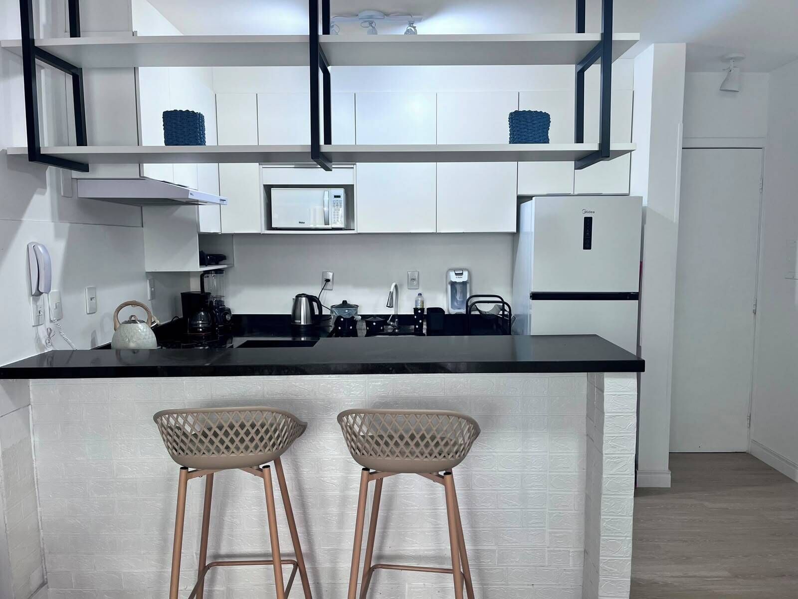 Een moderne keuken met witte kastjes, een zwart aanrechtblad, een ingebouwde magnetron en een koelkast. Er staan twee barkrukken met gevlochten zittingen.