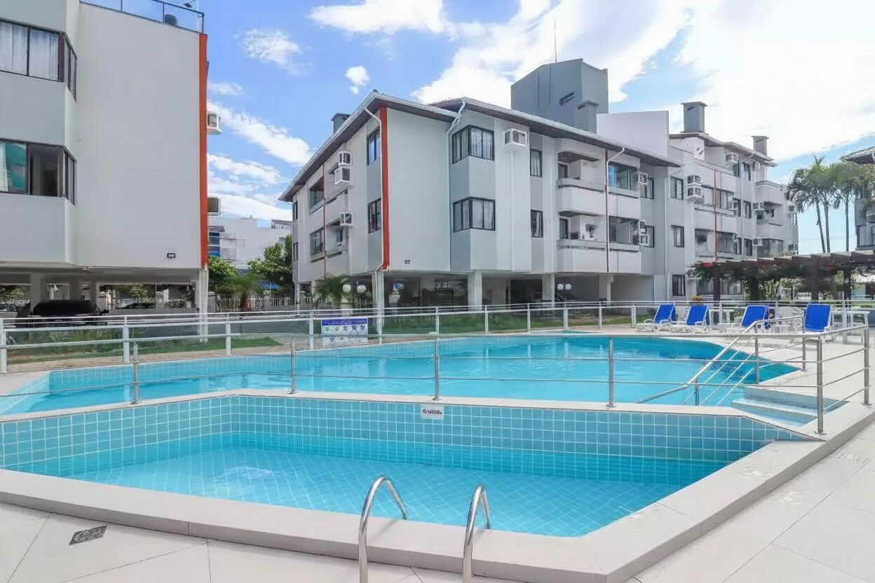 A rectangular swimming pool with a tiled deck, surrounded by a white fence, and adjacent to a multi-story building with balconies.