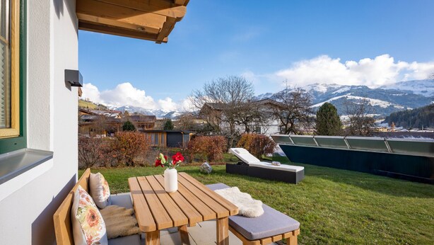 A wooden outdoor seating area with a table and benches, a vase with flowers, and a view of snow-capped mountains. - Horngacher Top 1 App (Kirchberg in Tirol)