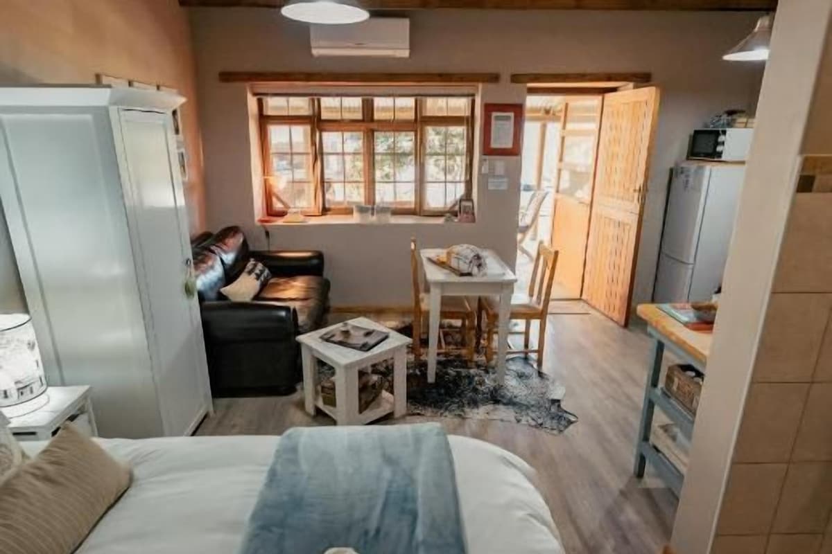 A bedroom with a bed, a bedside table, a window, and a kitchenette area.