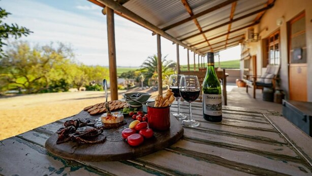 A covered outdoor dining area with a table set for a meal, including a bottle of wine, glasses, and a variety of food items. - Dassenheuwel Farm Cottages (Malmesbury)