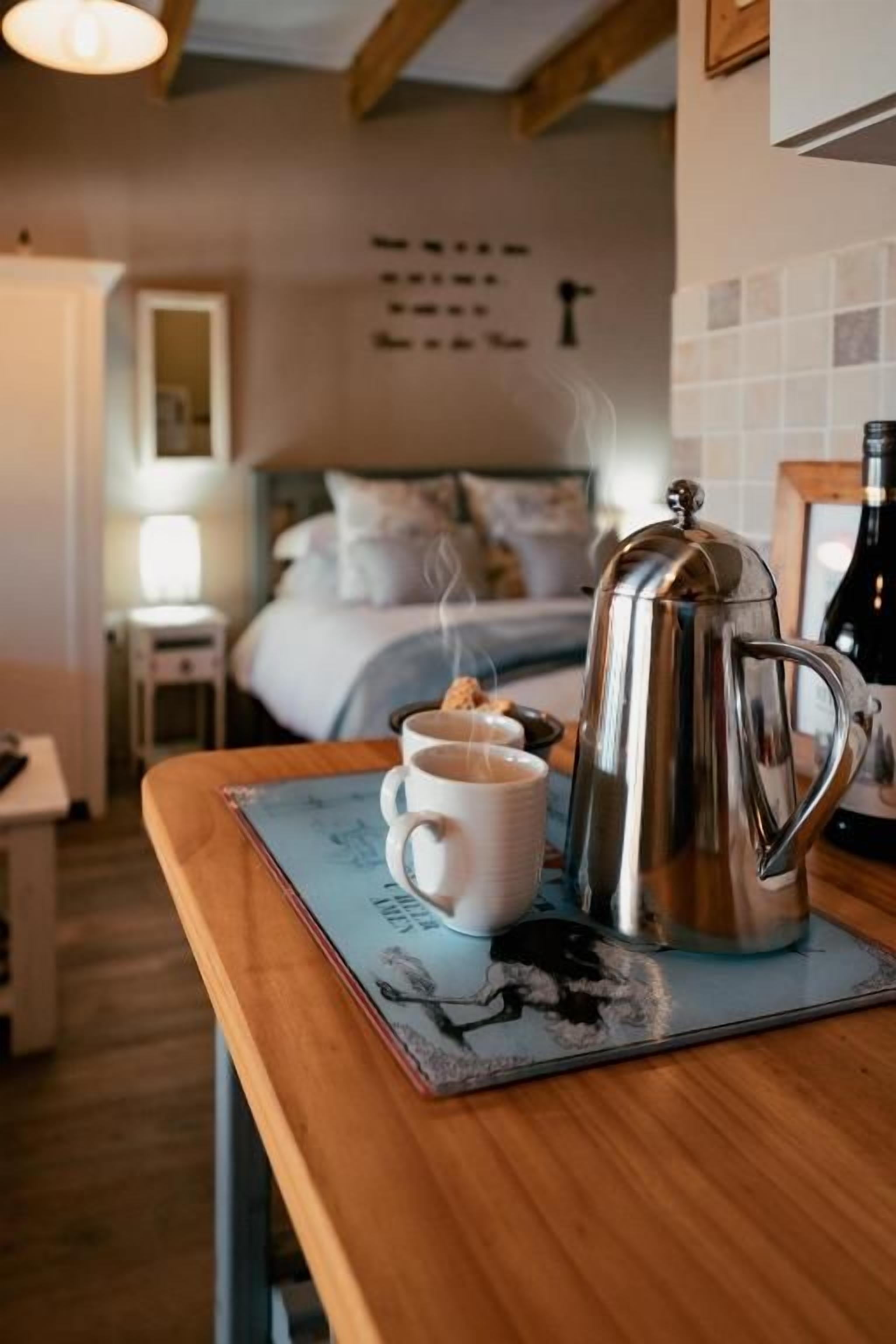 Una mesa de madera con una cafetera y una taza, una cama al fondo y una pared con un adorno.