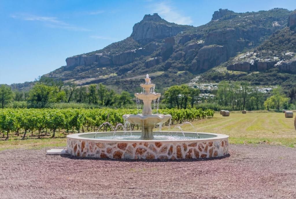 A circular stone fountain with water spouts in a landscaped area with vineyards and a mountain in the background.