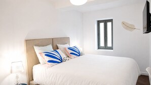 Un dormitorio con cama, mesita de noche, lámpara, ventana y televisor de pared.