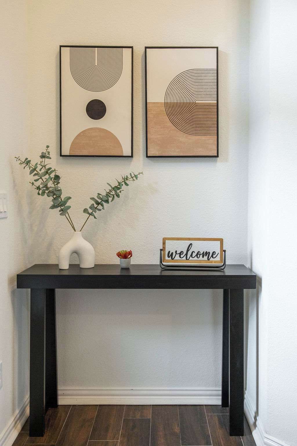 A black console table with a 'welcome' sign, two framed abstract artworks, and a vase with green foliage.