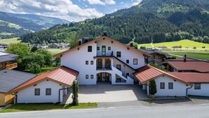 A large white building with multiple floors and a red roof, surrounded by lush green hills and a clear blue sky. - Horngacher Top 2 App (Kirchberg in Tirol)