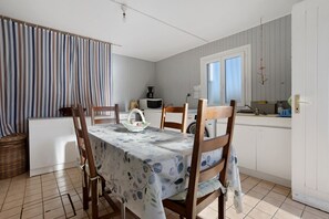 Comedor con mesa dispuesta para cuatro personas, sillas, zona de cocina con electrodomésticos y ventana con cortinas.