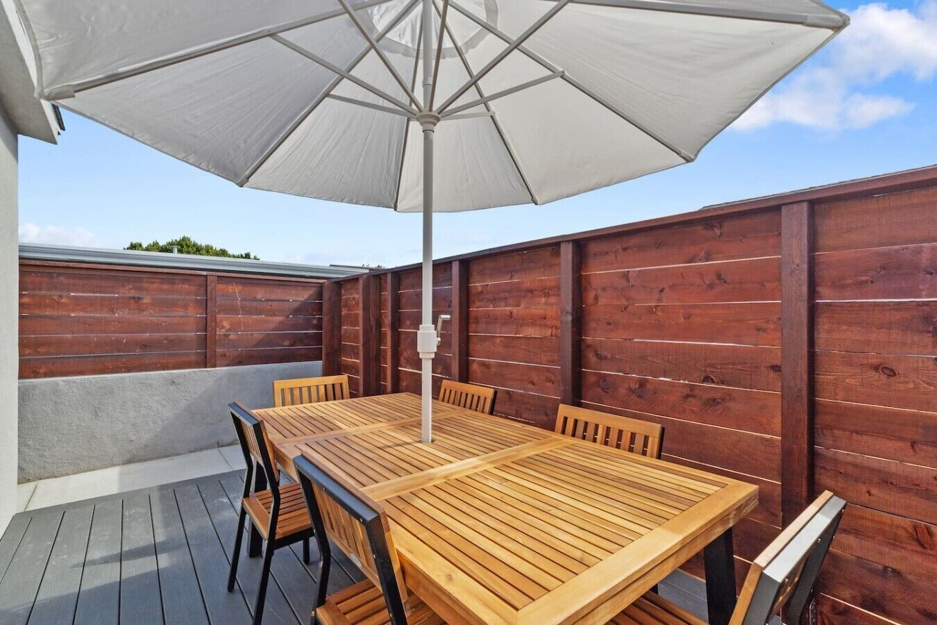 Um pátio externo com mesa e cadeiras de madeira, um guarda-sol branco e um cercado de madeira.