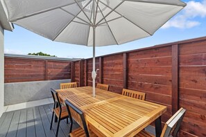 Eine Terrasse im Freien mit einem Holztisch, Stühlen, einem weißen Sonnenschirm und einem Holzzaun.