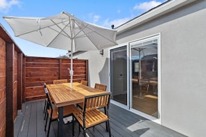 A wooden deck with a table and chairs, a large umbrella, and a sliding glass door leading to an indoor area.