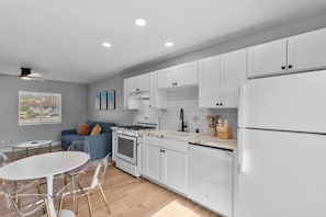 A modern kitchen with white cabinets, a stove, and a refrigerator. There is a dining area with a table and chairs. - Vista Home Base | Parking, Near Beaches & Shops (Vista)