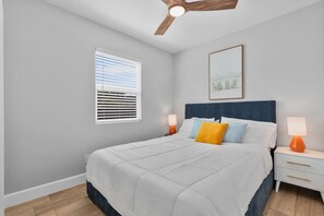 Ein Schlafzimmer mit einem Bett, Nachttischen, einem Deckenventilator, einem Fenster mit Jalousien und einem gerahmten Bild an der Wand.