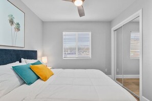 A bedroom with a bed, pillows, a ceiling fan, a window with blinds, and a door leading to another room.