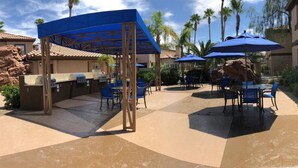 Un patio al aire libre con mobiliario cubierto, una parrilla de barbacoa y palmeras al fondo.