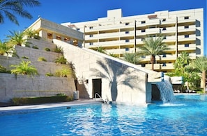 Une piscine agrémentée d’une cascade, entourée de palmiers et bordée d’un immeuble à plusieurs étages doté de balcons.
