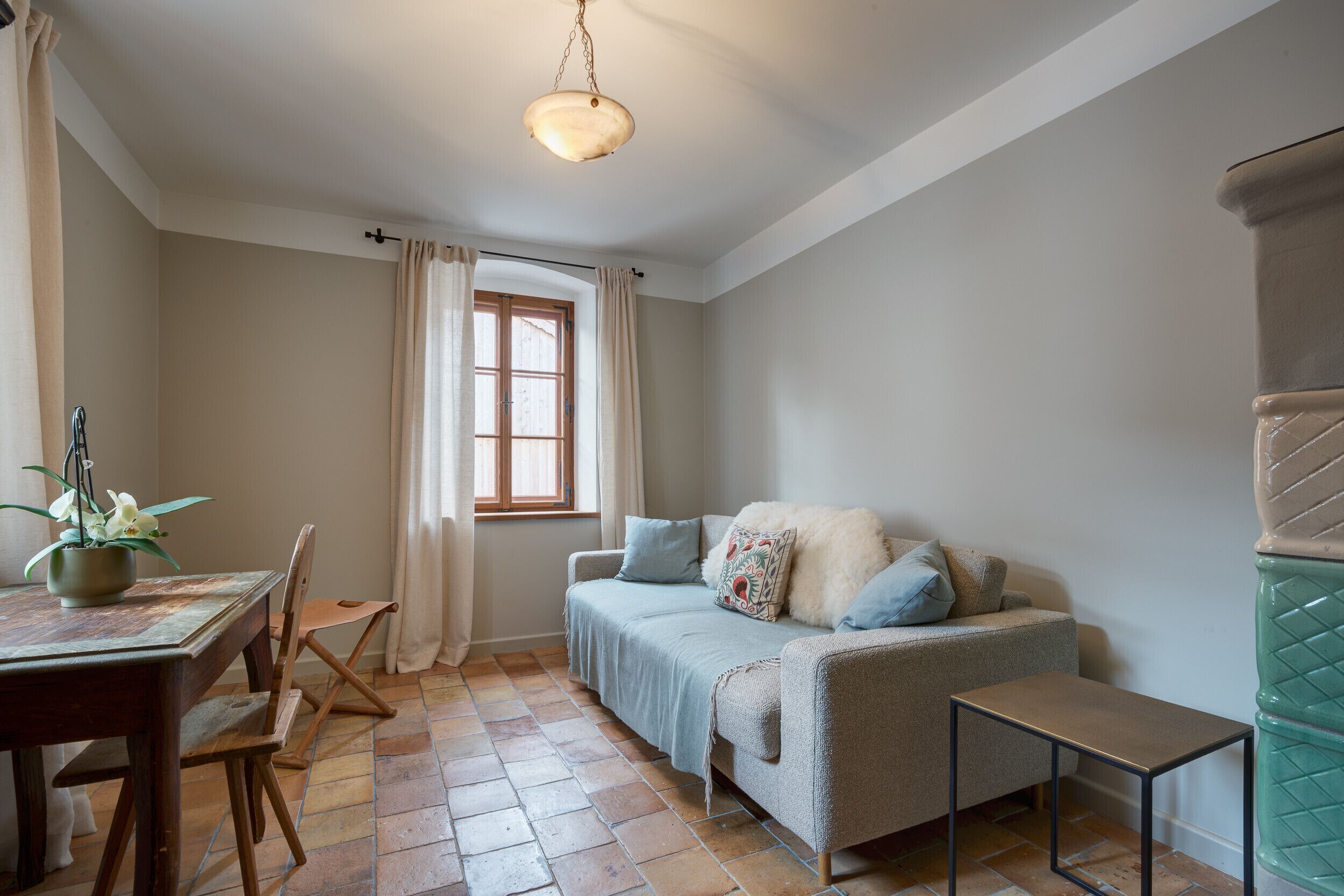 Una habitación acogedora con una mesa de madera, una silla, un sofá con cojines, una mesita auxiliar y una ventana con cortinas.