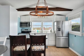 A modern kitchen with a dining area, featuring a hanging light fixture, stainless steel appliances, and a view through the window. - Cozy ocean front 3-bedroom cottage in beautiful Carolina Beach (Carolina Beach)