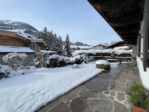 Ein verschneiter Innenhof mit Steinplatten, Holzbauten und schneebedeckten Bäumen.