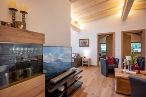 Ein modernes Wohnzimmer mit großem Flachbildfernseher, Holzmöbeln und Blick auf Berge durch das Fenster.