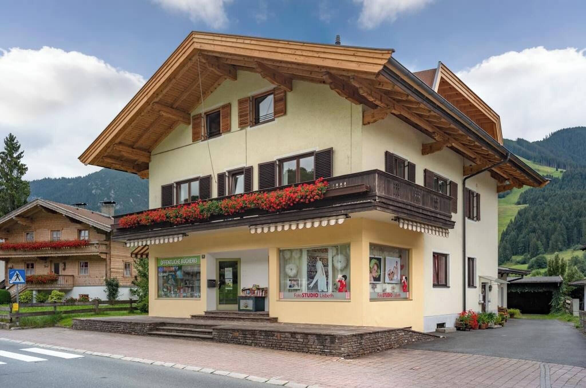 Un immeuble de deux étages doté d’un balcon orné de fleurs rouges, un commerce situé au rez-de-chaussée avec une enseigne, et un paysage montagneux en arrière-plan.