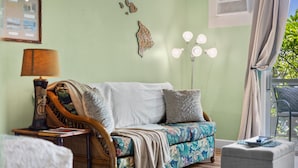 A room with a sofa, a side table, a lamp, a decorative wall piece, and a window with curtains. - Kailua Kona Islander | Steps to Beach, Lanai, Pool (Kailua-Kona)