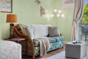 A room with a sofa, a side table, a lamp, a decorative wall piece, and a window with curtains. - Kailua Kona Islander | Steps to Beach, Lanai, Pool (Kailua-Kona)