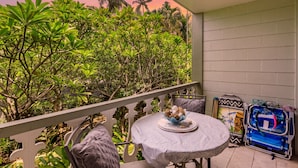 A balcony with a round table, chairs, and a view of dense greenery. - Kailua Kona Islander | Steps to Beach, Lanai, Pool (Kailua-Kona)