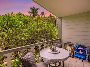 A balcony with a round table, chairs, and a view of dense greenery. - Kailua Kona Islander | Steps to Beach, Lanai, Pool (Kailua-Kona)