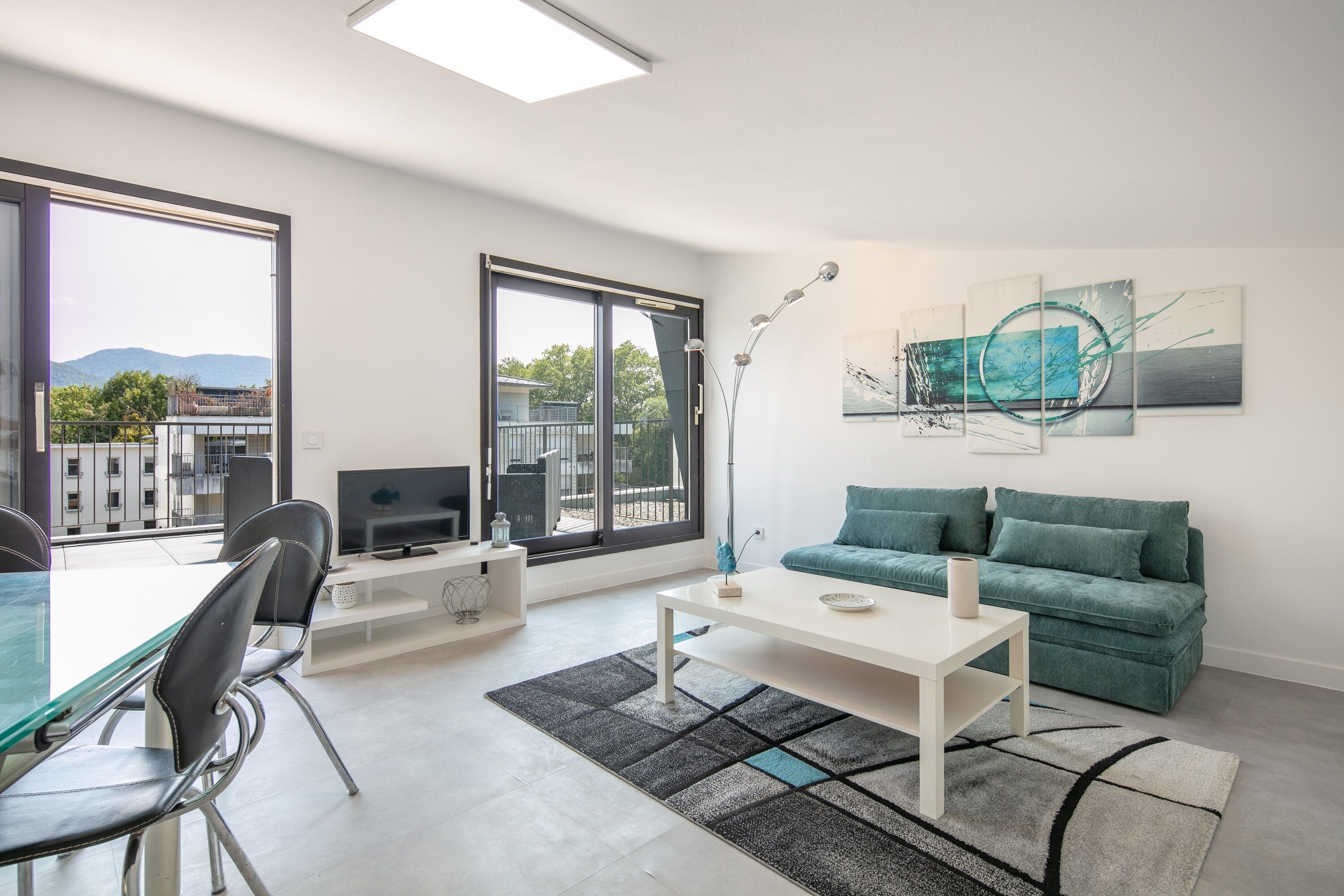 A modern living room with a sofa, a coffee table, a TV, and a large window offering a view of the outdoors.